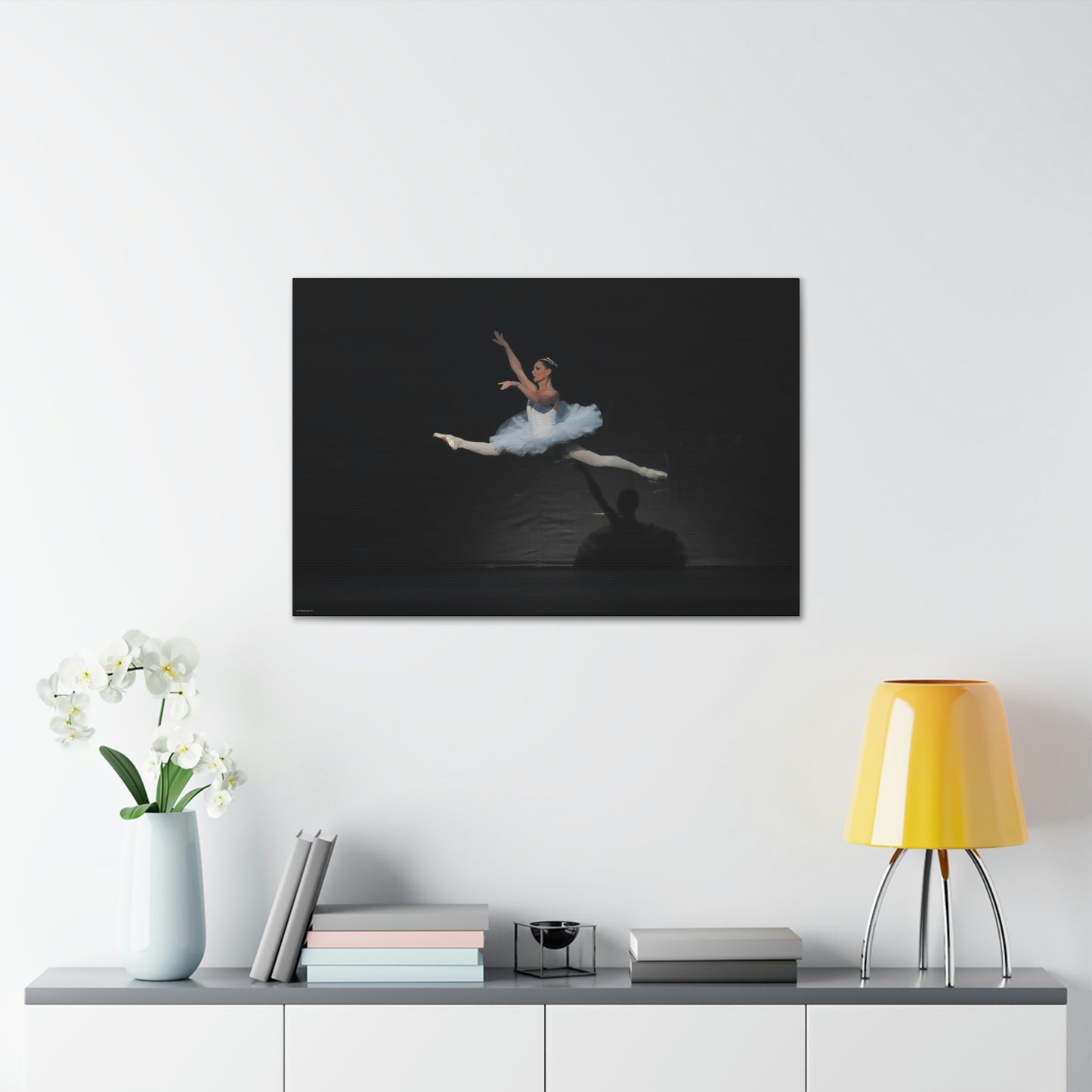 Canvas print on a white wall over a desk of a ballerina in a white tutu performing on stage in a mid-air jump with light casting her shadodw on a black backdrop.