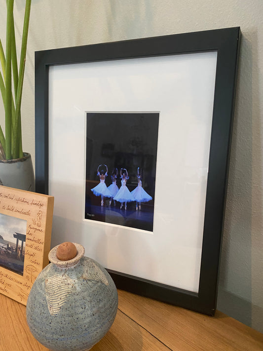 photograph of a painting in a black frame and white matte of four graceful ballerinas performing on a stage in white dresses with a dark backdrop