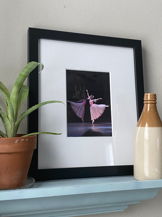 a photo of a painting framed in black with a white matte of two Graceful ballet dancers performing on stage in pink on a shelf in a home setting