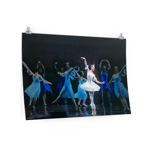 Prima ballerina dressed in white performing on a stage with a black backdrop and girls dancing around her in dark and light blue costumes